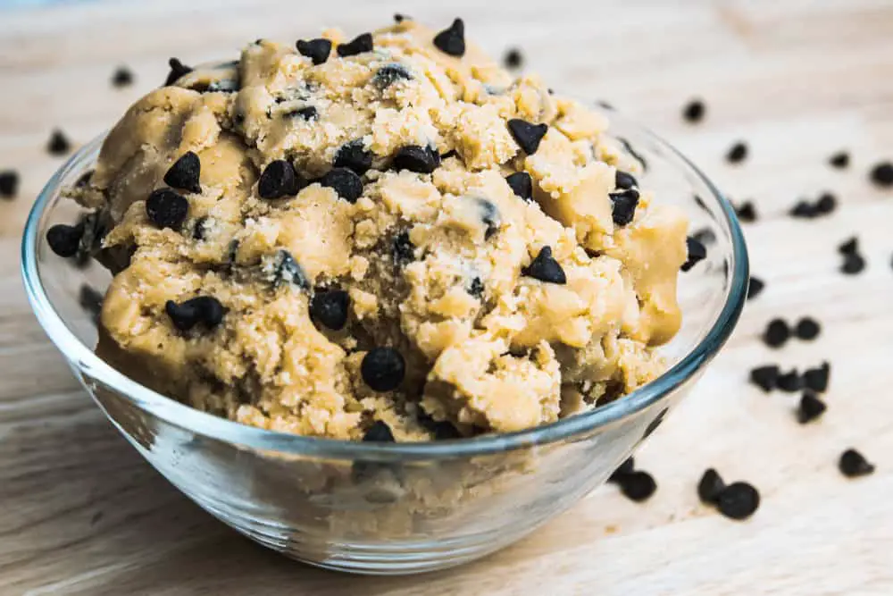 Glass bowl full of chocolate chip cookie dough with chocolate chips spread across counter