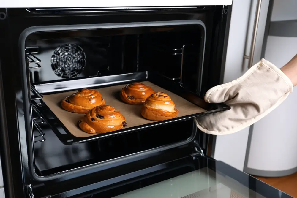 Hand wearing oven mitt pulling tray of freshly baked rolls out of the oven; one of the best ovens for baking